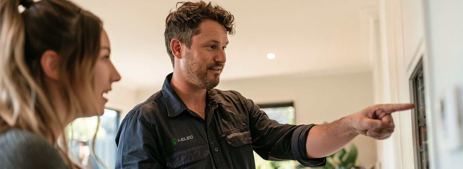 a licensed i-ELEC Australian electrician in a neat uniform explaining home electrical protection to a homeowner in a modern Sydney house, electrician gesturing towards a wall-mounted electrical area in the background