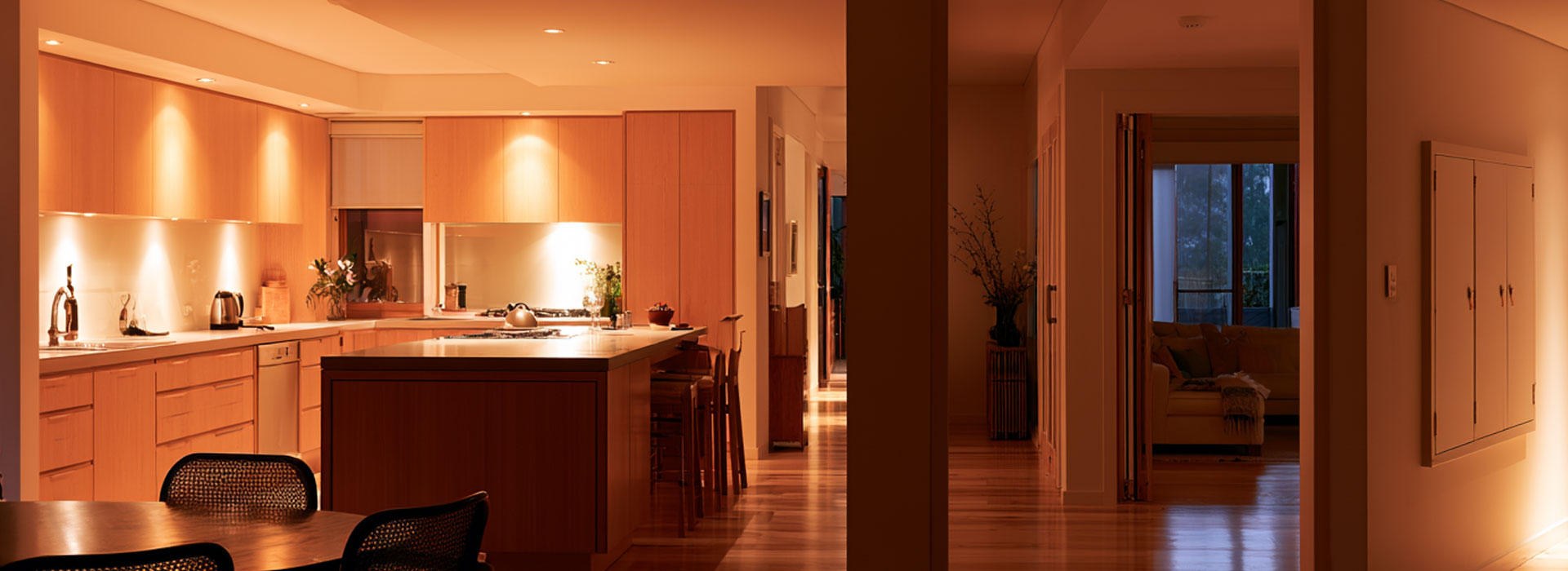 contemporary Sydney home interior at dusk, warm architectural lighting throughout a clean open-plan kitchen and living area, calm safe atmosphere, subtle closed electrical switchboard cabinet in the hallway