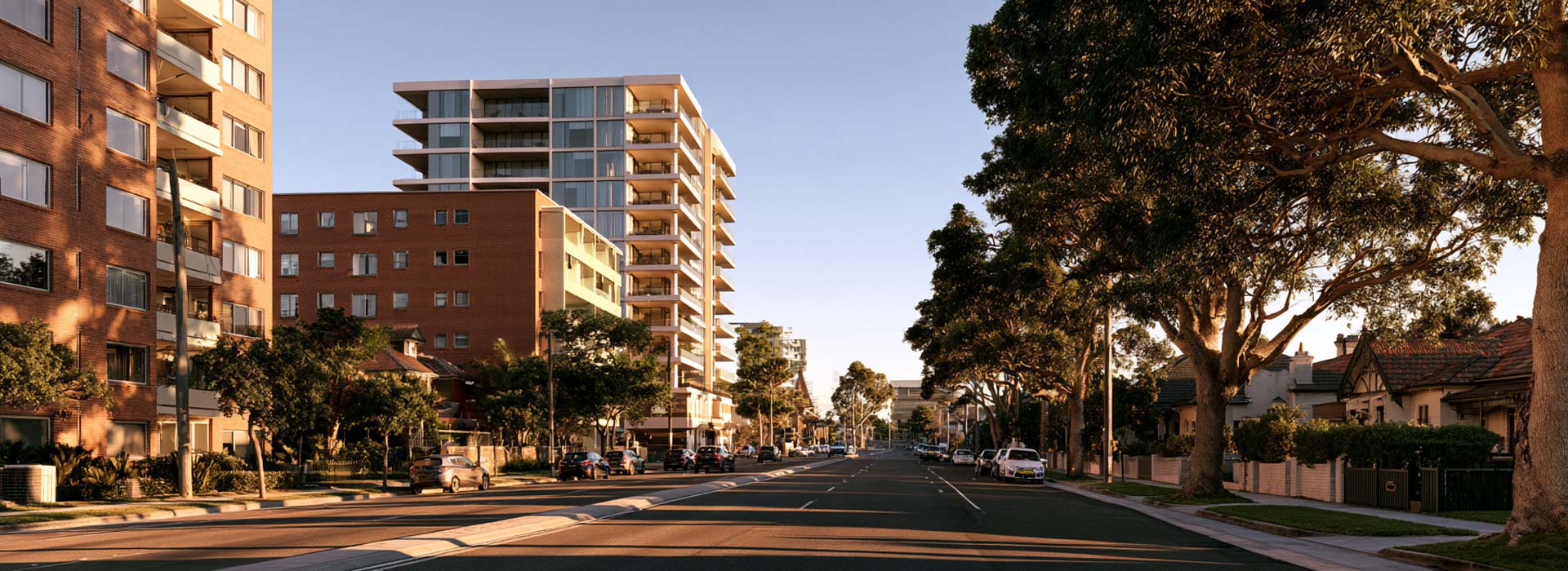 streetscape in Kogarah, Sydney, modern mid-rise apartment buildings mixed with older red-brick unit blocks and established detached Australian homes, subtle St George Hospital precinct visible in the distance