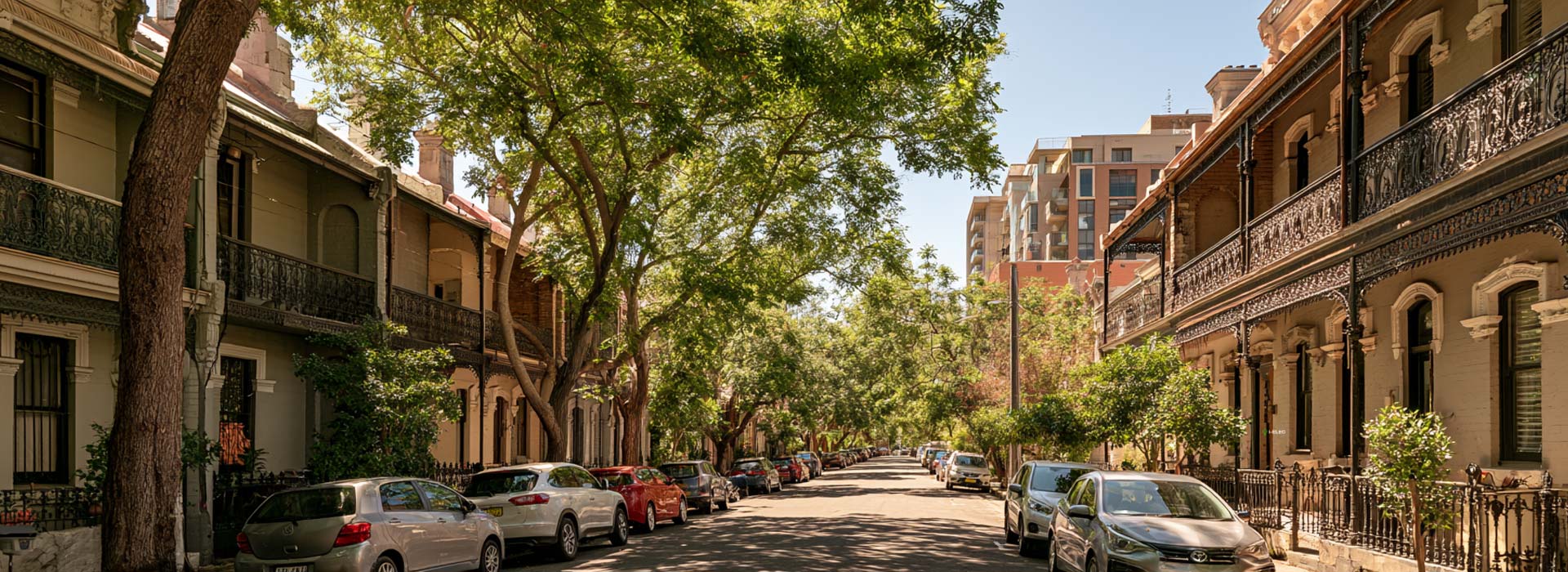 wide photograph of a Redfern Sydney residential street, restored two-storey Victorian terrace houses with ornate wrought-iron balconies, mature street trees, Australian parked cars, subtle modern apartment buildings in the distance, clear summer daylight