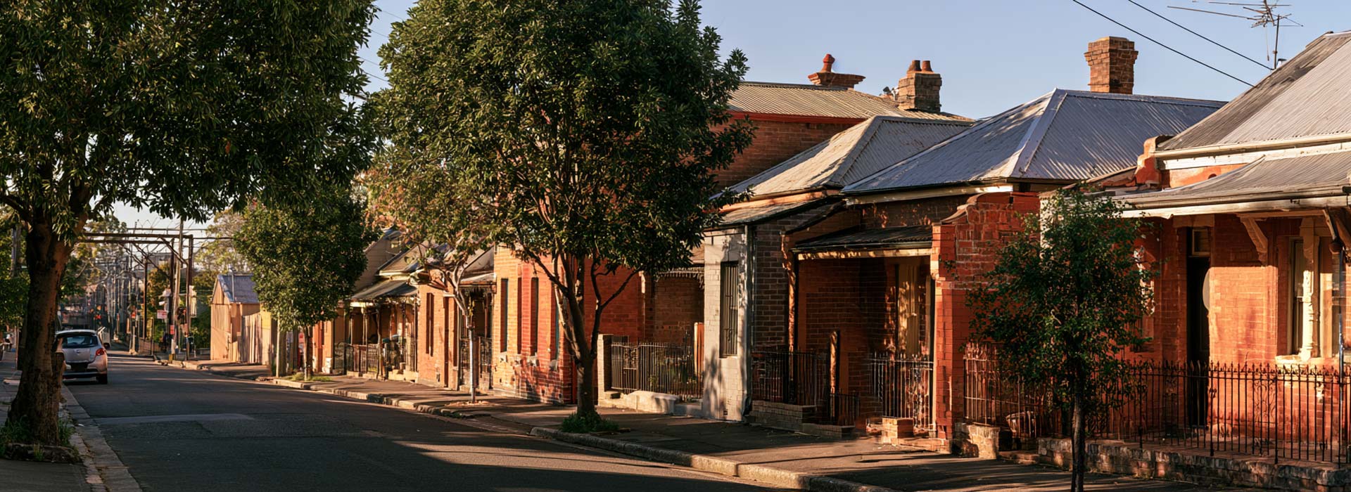 panoramic photograph of a Sydenham Sydney residential street, compact Federation-era red brick cottages and terraces with corrugated metal roofs, mature street trees, subtle railway gantries visible in the distance